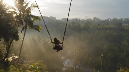 travel woman swinging over jungle at sunrise enjoying exotic vacation sitting on swing with sun flare shining through palm trees in tropical rainforest holiday lifestyle freedom