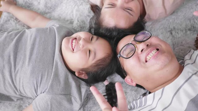 Top view of happy Asian family lying on the floor in living room, looking at camera and making two fingers pose. Staying with family at home in the weekend. Happy family concept