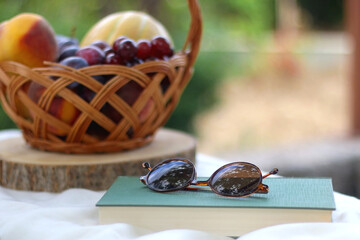 Vintage basket full of various fruit, hardback book and retro sunglasses on picnic basket. Selective focus.