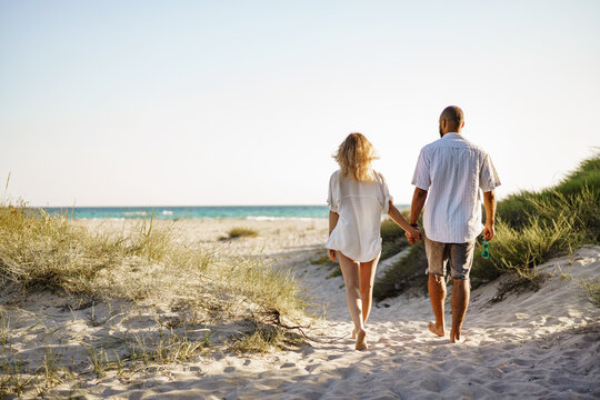 Young happy couple holding hands and walking together to the beach on summer day