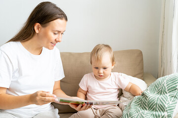 Horizontal of mummy reading book with curious adorable blond child in casual clothes while sitting on sofa in light living room. Concept of parenthood, child education and development. Love and care