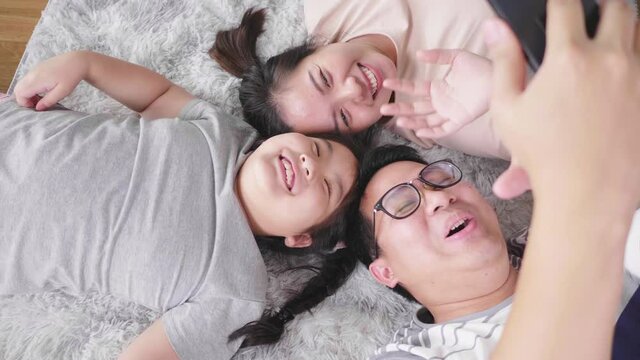 Top view of happy Asian family lying on the floor in living room, taking video call and making waving hands. Staying with family at home in the weekend. Happy family concept