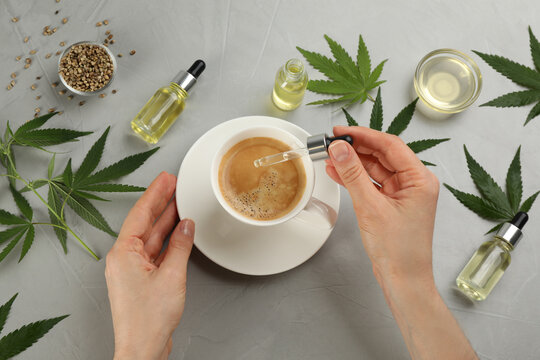 Top view of woman dripping THC tincture or CBD oil into coffee at light grey table, closeup