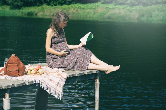 Pregnant woman sitting on wooden pier near lake and reading book. Outdoor picnic near water. Wellbeing and time to relax alone.