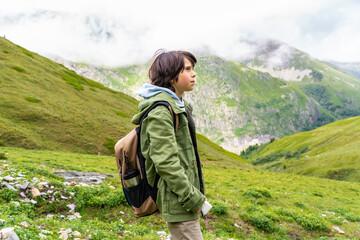 Naklejka na ściany i meble Portrait of a young boy with backpack in a green jacket standing in the mountains in French Alps with his head up, looking away with a serious face. Hiking with children.
