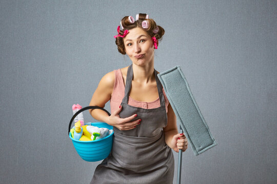 Comical female washer wear hair rollers and apron holding mop. Funny housewife cleaning around with a mop on gray background.