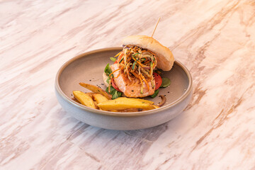 Fish burger with a Norwegian salmon taco with stewed vegetables, tomato and lamb's lettuce garnished with French fries on a gray tray and marble table