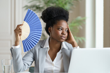 Tired young african businesswoman hold wave fan exhausted of heat indoors overheated at workplace...