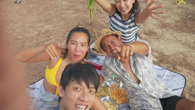 Group of Asian man and woman friends sitting on the beach and using smartphone taking selfie or video call together on summer vacation. Male and female friendship having fun outdoor lifestyle activity