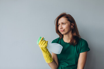 Dissatisfied young woman holding spray detergent while wearing yellow rubber gloves for hands protection during cleaning isolated over grey background copyspace 