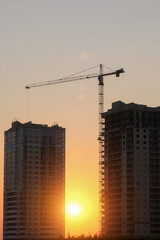 Fototapeta na wymiar Against the backdrop of the setting sun, there are two houses under construction and a construction crane. Vertical photo.