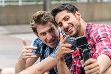 Two blogger handsome friends taking selfie together with action camera sitting on street city looking the camera making enjoyment face. Two happiness traveler man take picture while travel in holiday.