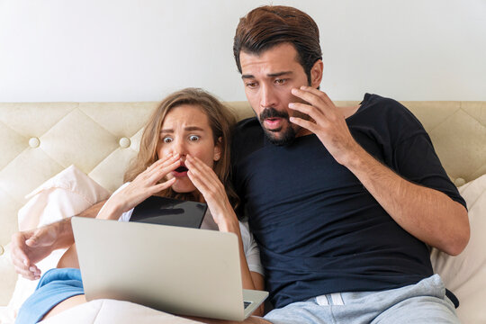 Scared young couple sitting under blanket on the bed with laptop while watching horror movie at tv together in the bedroom. Husband embarrassed his wife while looking scary shocked at home.