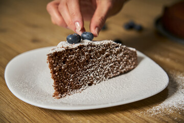 A woman with a passion for cooking decorates a slice of handmade chocolate cake in her home kitchen with blackberries, red currants and a sweet white rain of powdered sugar.