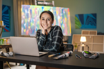 Portrait of smiling brunette wearing a checkered shirt sitting at a desk in front of a laptop in...
