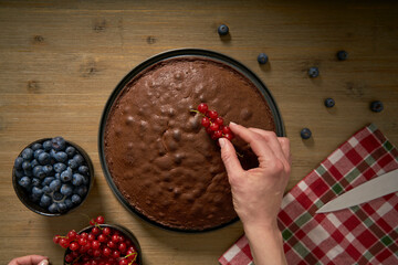 Woman with a passion for cooking decorates handmade chocolate cake in her home kitchen with blackberries and red currants.