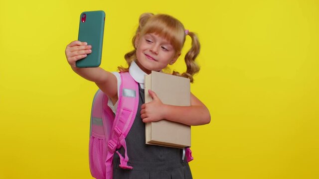 Blond teenage student girl kid blogger taking selfie on mobile phone for social media isolated on yellow studio background. Education study, back to school. Child in school uniform wears pink backpack