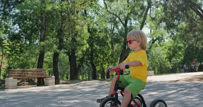 The boy rides a tricycle in the park, the camera zooms in on the boy.