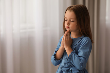 Fototapeta na wymiar Cute little girl with hands clasped together praying indoors. Space for text