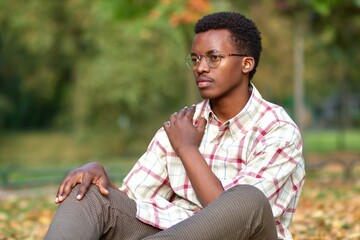Naklejka na ściany i meble Portrait of serious pensive thoughtful unhappy sad guy, young black African Afro American ethnic man in glasses and shirt is sitting on ground on leaves in golden autumn, fall park. 