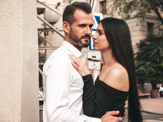 Beautiful fashion woman and her handsome elegant boyfriend in suit. Sexy brunette model in black evening dress. Fashionable couple posing in the street in Europe. Brutal man and his female outdoors