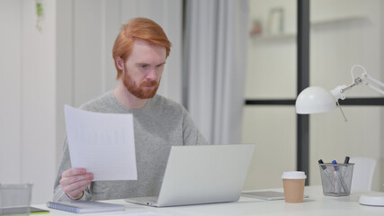 Beard Redhead Man Reading Paper While using Laptop at Work 