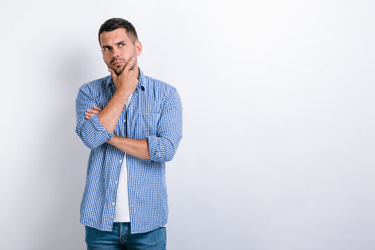 Portrait of pensive handsome bearded young man standing, touching his face, looking aside away and thinking about something. Indoor studio shot, isolated on white background