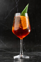 Glass of ice cold Aperol spritz cocktail served in a wine glass, decorated with slices of orange and basil leaf, placed on a black background