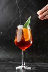 Glass of ice cold Aperol spritz cocktail served in a wine glass, decorated with slices of orange and basil leaf, placed on a black background