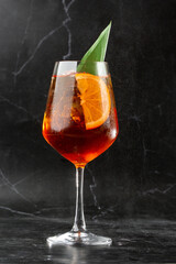 Glass of ice cold Aperol spritz cocktail served in a wine glass, decorated with slices of orange and basil leaf, placed on a black background
