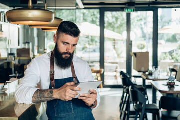 Handsome Barista Using Digital Tablet in a Cafe.
Serious waiter with a beard leaning on the bar counter and reading online order or watching something on a digital tablet while working in coffee shop.