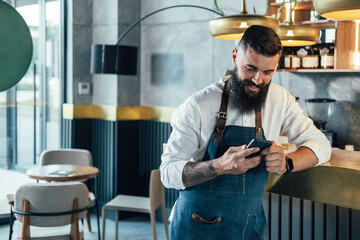 Happy Barista Using Mobile Phone in a Cafe.
 
Cheerful smiling waiter with a beard leaning on the...