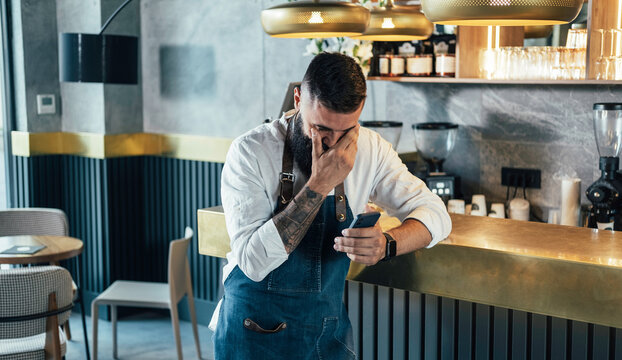 Happy Barista Using Mobile Phone in a Cafe. 

Cheerful smiling waiter with a beard leaning on the bar counter and crying while looking something on his smartphone in the coffee shop. 