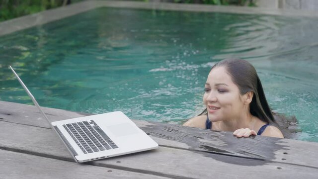 A beautiful mixed-race girl emerges from the pool and answers a video call on her laptop. a female freelance programmer is talking via video communication with colleagues at work. Slow motion.