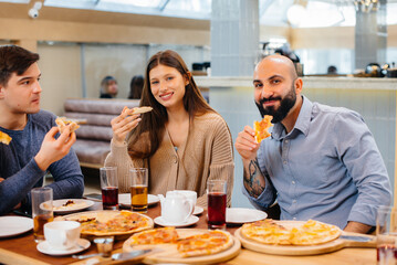 A group of young cheerful friends is sitting in a cafe talking and eating pizza. Lunch at the pizzeria.
