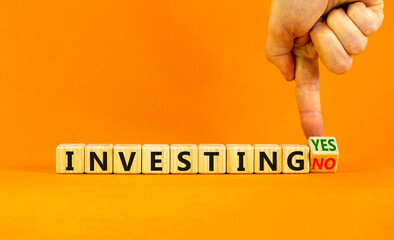 Investing yes or no symbol. Businessman turns a wooden cube and change words 'investing no' to 'investing yes'. Beautiful orange background. Business and investing yes or no concept, copy space.
