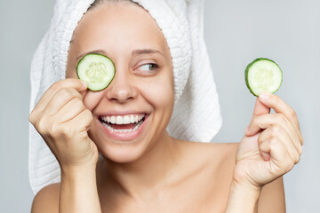 A young pretty happy smiling woman with a white towel on her head after a shower holds cucumber slices in her hands to make a refreshing face mask isolated on a gray background. Skin care, cosmetology - Powered by Adobe