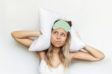 A young dissatisfied blonde woman in a green sleep mask covers her ears with a pillow looking up isolated on a white background. Noisy neighbors from above prevent the girl from sleeping