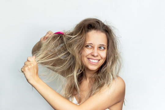 A young blonde woman combing her tangled unruly dry bleached hair with a pink comb isolated on a white background. Hair care