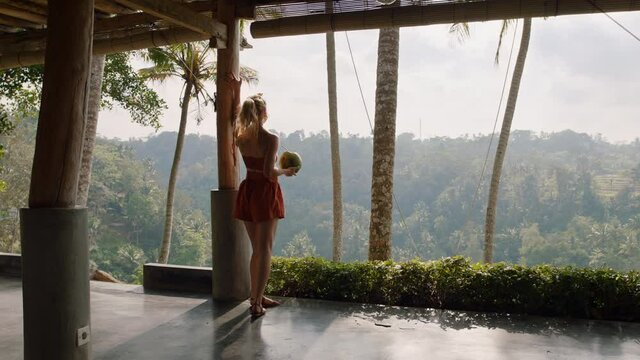 young tourist woman drinking coconut cocktail at tropical hotel resort looking beautiful view of jungle enjoying exotic vacation lifestyle in paradise