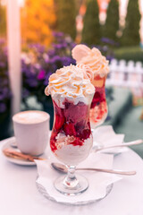 Strawberry ice cream dessert with whipped cream and crushed meringue, served in a glass cup. A cup of coffee on the coffee table. Purple flowers in the background, Darłowo Poland