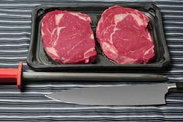 Fresh rib eye steak, Meat and food industry. Juicy premium cut of beef. Quality product on black plastic tray on black and white apron, knife and sharpening steel. Butcher craft