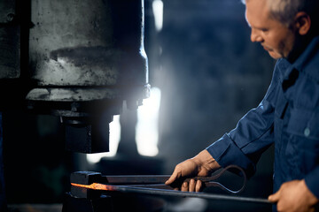 Caucasian man in blue overalls bending molten metal under press machine at forge. Blacksmith...