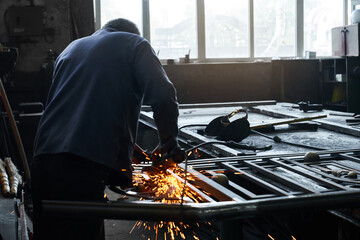 Craftsman in uniform using angle grinder for polishing metal at workshop garage. Competent worker making sparks flying around. Processing steel. 