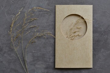 Dry grass with a mockup of kraft paper card  with a cut-out circle on gray old plaster.  Stylish greeting card, invitation.