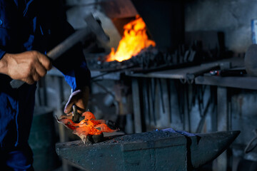 Industrial worker in blue overalls forging molten metal on anvil. Close up of skillful man creating a handmade product at forge. Working process. 