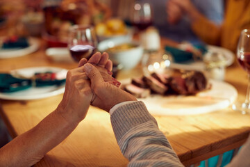Close-up of senior couple holds hands while blessing food during Thanksgiving meal with family at dining table.