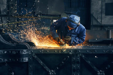 Competent industrial worker dressed in protective clothes, glasses and gloves grinding metal construction using polishing machine. Sparks from metal processing. - obrazy, fototapety, plakaty
