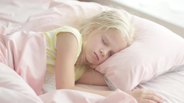 Adorable blonde girl sleeping sweetly in the morning on pink bed linens at home. Childrens dreams, comfort, rest and peace.