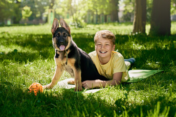 Smiling little boy lying on grass with german shepherd puppy. Happy owner playing with lovely dog...